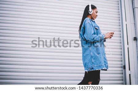 Positive hipster girl in earphones listening audio record with high volume and sending text message on mobile app, millennial woman in jeans jacket walking and enjoying music on radio via headphones