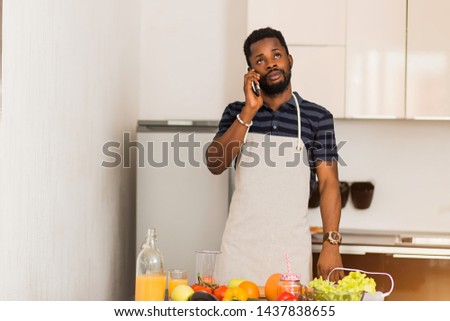 Handsome black guy wearing apron talking on the mobile phone while cooking, has thoughtful expression, does not know what to prepare, standing in kitchen at home.