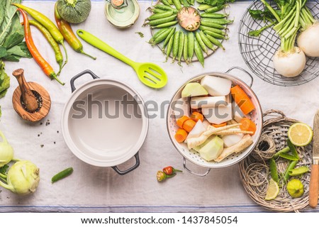 Cooking pot with spoon and white colander with fresh vegetables for vegetarian stock , broth or stew  in light kitchen table background, top view.  Copy space. Healthy food eating