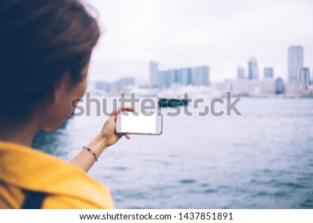 Back view of female influencer photographing Victoria bay for share content to social media web page, millennial woman tourist making images via cellular with blank screen during online blogging