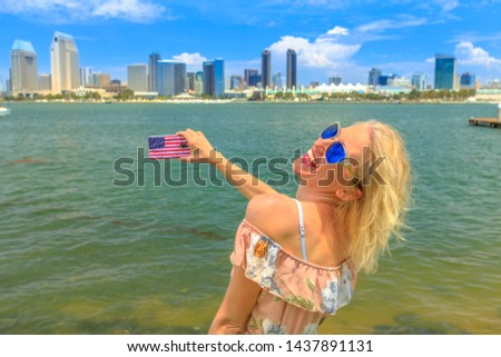 Blonde happy tourist takes photo of San Diego By skyline by mobile phone with American flag cover. Woman lifestyle takes picture in California summer holidays with smartphone from Coronado Island, USA