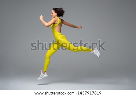 Excited young lady in yellow sportswear smiling and leaping while exercising against gray background