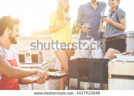 Group of young friends having fun at patio barbecue party drinking wine and laughing - Happy chef making fire with bellows for bbq dinner - Focus on left man hands - Youth, food, friendship concept