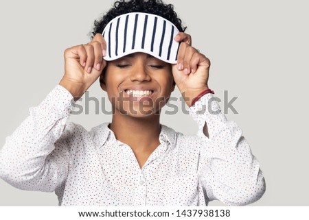 Smiling african American millennial woman isolated on grey studio background wear sleeping stripy eye mask, happy biracial young girl with sleepwear accessory on head feel healthy refreshed after nap