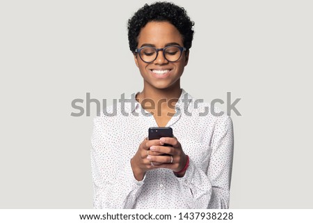 Smiling african American girl wear glasses isolated on grey studio background hold smartphone chatting or texting, happy ethnic biracial woman use cellphone read good news, browsing shopping online Royalty-Free Stock Photo #1437938228