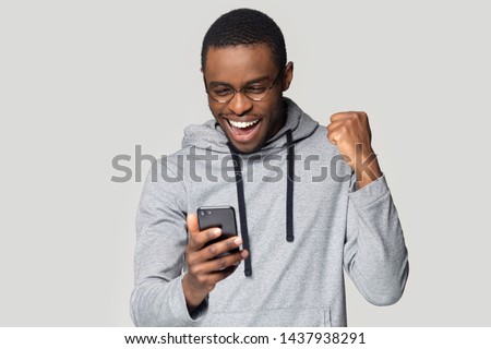 Excited african American millennial male in glasses isolated on grey studio background hold smartphone happy wining online, overjoyed black man look at cellphone smile feel joyful reading good news Royalty-Free Stock Photo #1437938291