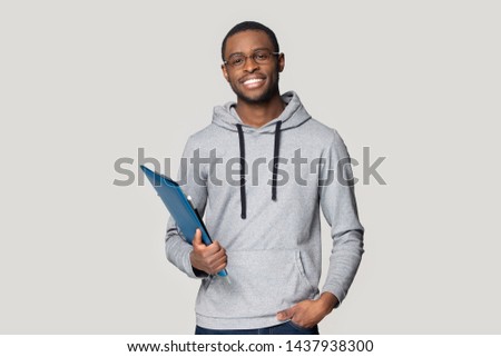 Smiling african American man in glasses isolated on grey studio background look at camera keep documents in hand, happy black male student or employee stand holding binder case and gadget