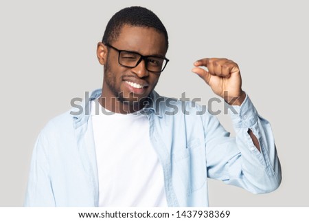 Smiling millennial african American male in glasses isolated on grey studio background show small prices with finger gesture, happy black man look at camera use hand demonstrate little measurement