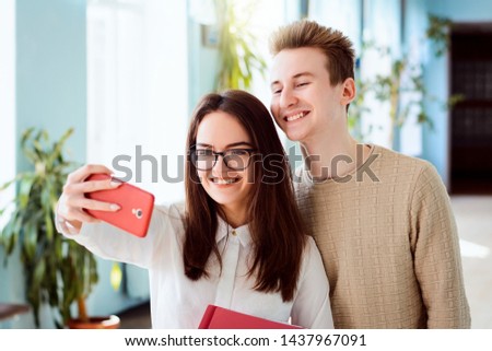 Couple of young cheerful students making selfie on red mobile phone standing indoor in university campus holding books. Happy students having good time together after classes