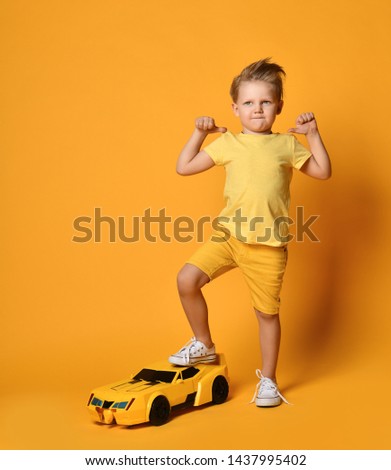 Kid boy in yellow t-shirt and shorts thumbs up his leg steps on a big race car as birthday present happy thinking something over on yellow background
