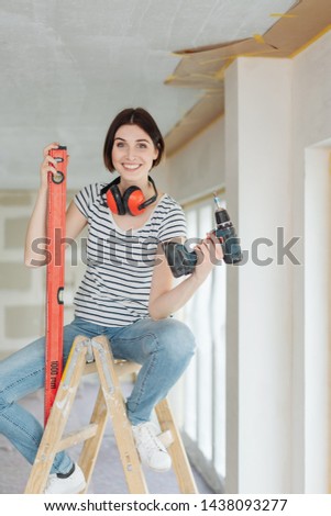 Happy young woman doing DIY home improvements seated on a wooden ladder holding a builders level and electric drill smiling at the camera