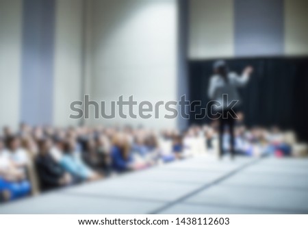 Woman speaker and audience at conference.
Blurred female presenter during meeting. Stage at business seminar. Lecturer giving speech photo. People attend workshop.
