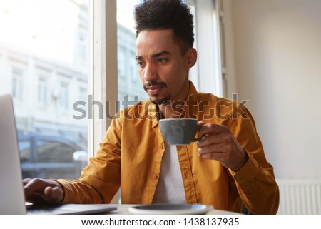 Portrait of young attractive African American boy, works at a laptop in a cafe, drinks coffee and thoughtfully looks at monitor, concentrate on his work.