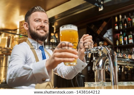 Positive, handsome barman holding cold lager beer glass. Cheerful brewery worker standing at bar counter, smiling. Adult man in white shirt and brown apron looking at camera. Royalty-Free Stock Photo #1438164212
