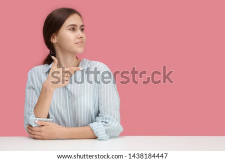 Horizontal shot of beautiful joyful young in stylish striped blue shirt working at office desk and making gesture, pointing index finger at blank pink wall with copyspace for your promotional content
