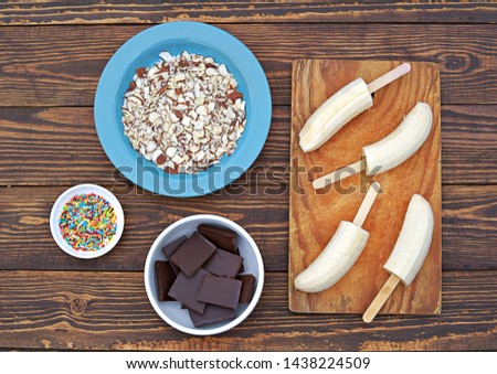 Ingredients for cooking frozen bananas in chocolate on wooden background: fresh bananas, chocolate, almond petals, sugar topping, sticks. Top view, copy space.