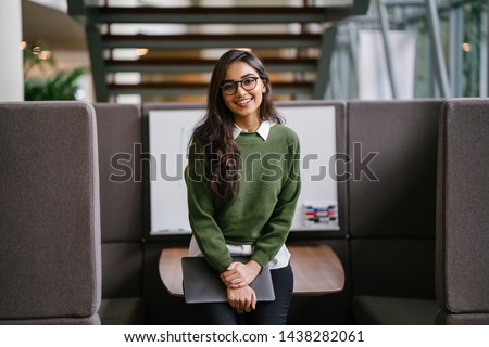 Portrait of a young, beautiful, intelligent and attractive Indian Asian MBA student smiling as she leans in a discussion booth in her campus. She is wearing a preppy outfit and smiling confidently.  Royalty-Free Stock Photo #1438282061