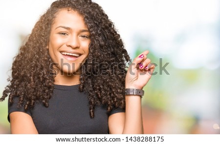 Young beautiful woman with curly hair with a big smile on face, pointing with hand and finger to the side looking at the camera.