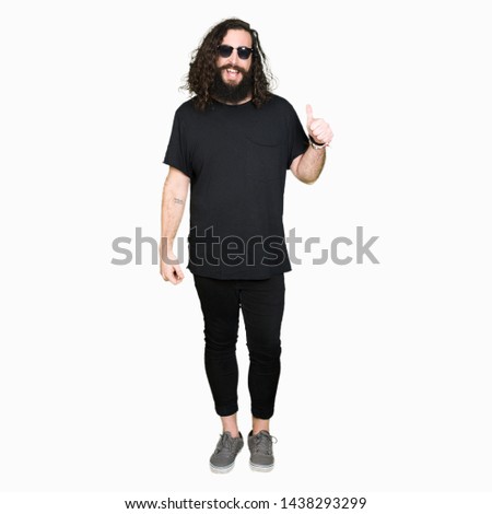 Young hipster man with long hair and beard wearing sunglasses doing happy thumbs up gesture with hand. Approving expression looking at the camera with showing success.