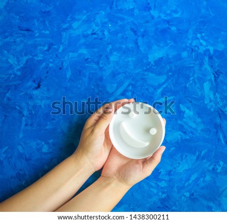 Smiley white face in the hands of a woman on a blue background with copy space