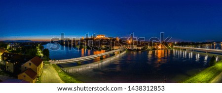 Castle on top of hill at night, aerial view of Ptuj, Slovenia in dusk, illuminated bridge across a river, reflection in the water, ancient Roman old town