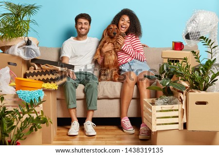 Relocation, renovation and removal concept. Happy diverse couple sit on sofa with favourite dog, have break on moving day, busy unpacking, have mess in new flat, many packages, remove to own house Royalty-Free Stock Photo #1438319135