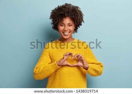 Be my valnetine. Charming young girlfriend confesses in love, shows heart gesture, smiles broadly, has romantic feelings, seeks lonely hearts, feels passion, dressed in casual bright yellow sweater