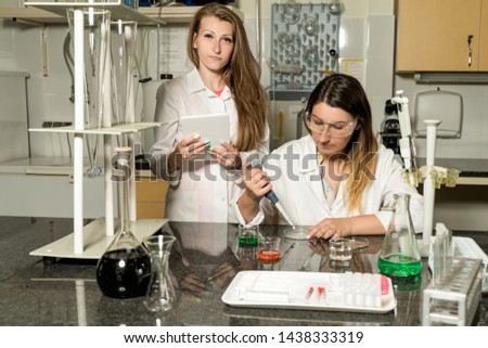 Team of two, Caucasian white, female laboratory technicians in white robes working in chemical or pharmaceutical laboratory, laboratory equipment and glassware in background