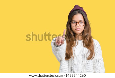 Young beautiful brunette hipster woman wearing glasses and winter hat over isolated background Pointing with finger up and angry expression