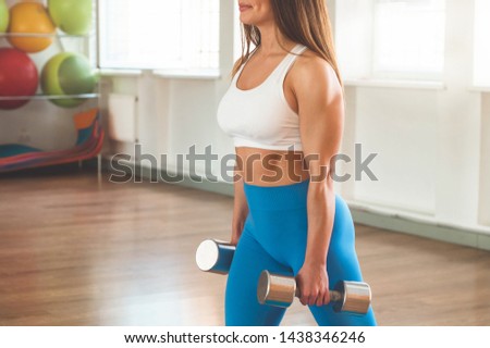 Fitness woman holding dumbbells. Fitness instructor in the sport room background. Female model with muscular fit and slim body.
