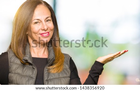 Beautiful middle age woman wearing winter vest smiling cheerful presenting and pointing with palm of hand looking at the camera.