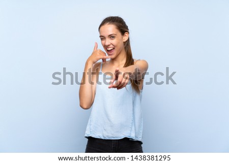 Young woman over isolated blue background making phone gesture and pointing front