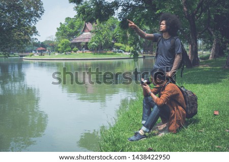 Two male Asian travelers relax by sitting on green grass near pond in public park, they looking around garden and take a photo from camera. copy space for text