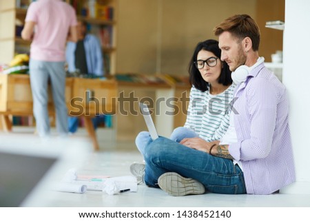 Busy young colleagues in casual clothing sitting on floor in coworking space and using laptop while analyzing internet resources together