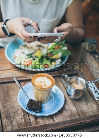 Woman's hands taking photo of coffee cup, Piccolo Latte, brownies with breakfast on wooden table by smartphone vertical style.