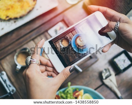 Woman's hands taking photo of coffee cup, Piccolo Latte, brownies with breakfast on wooden table by smartphone, top view.
