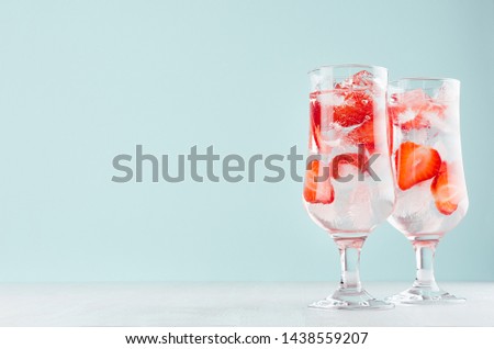 Fresh fitness strawberry mineral water with ripe strawberry slices, ice cubes and tonic in elegant glass on white wood table, mint color wall.