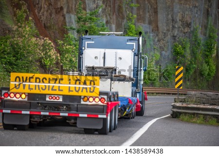 Big rig powerful heavy-duty semi truck tractor transporting oversized empty step down semi trailer with oversize load sign driving on the mountain road with rock wall