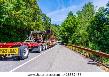 Big rig powerful heavy-duty semi truck tractor transporting oversized excavator on step down semi trailer with oversize load sign driving on the forest road with green trees