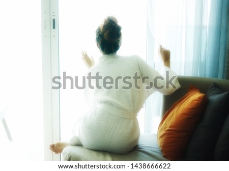 Soft photo of woman in bathrobe sitting next to curtain.