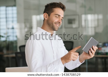 Young businessman smiling while standing alone in a large modern office working online with a digital tablet