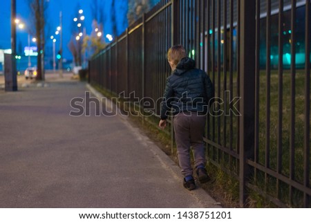 A little boy walks along a metal fence in the evening.