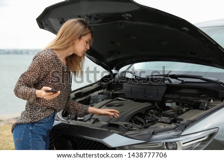 Troubled young woman with phone near broken car outdoors