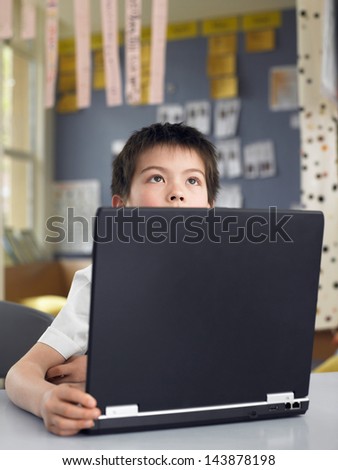 Thoughtful elementary schoolboy sitting by laptop in classroom