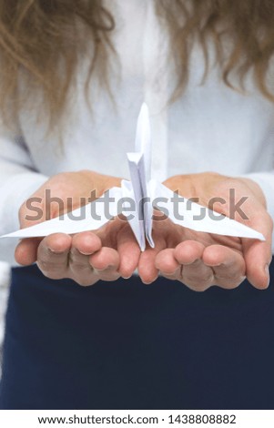 Female hands holding white paper crane on blurred background with young girl wearing a white shirt and blue skirt