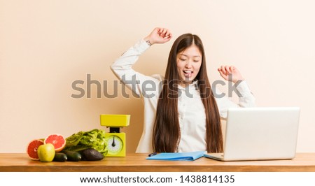 Young nutritionist chinese woman working with her laptop celebrating a special day, jumps and raising arms with energy.