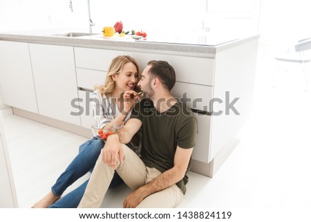 Photo of a smiling cute young loving couple indoors at the kitchen eat vegetable salad have a breakfast sitting on floor.