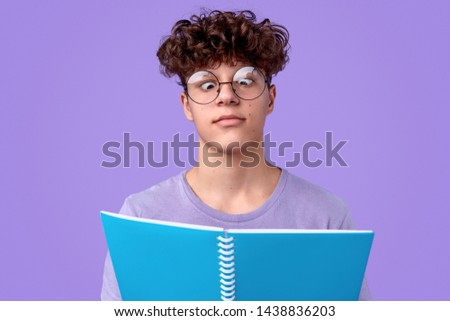 Nerdy youngster in spectacles crossing eyes while reading notes in notepad against violet background