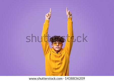 Stylish youngster in yellow hoodie and sunglasses looking a camera and pointing up with both hands against violet background