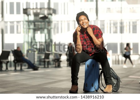 Portrait of happy younga travel man sitting on suitcase while talking on mobile phone in airport